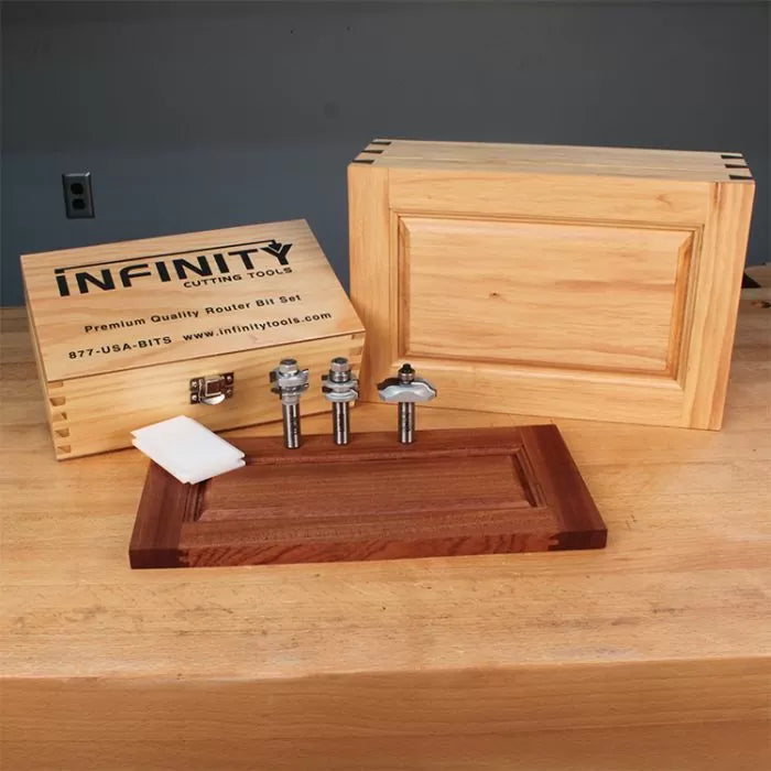 Router bit set with three bits, wooden storage box, and cabinet door panels on workbench.