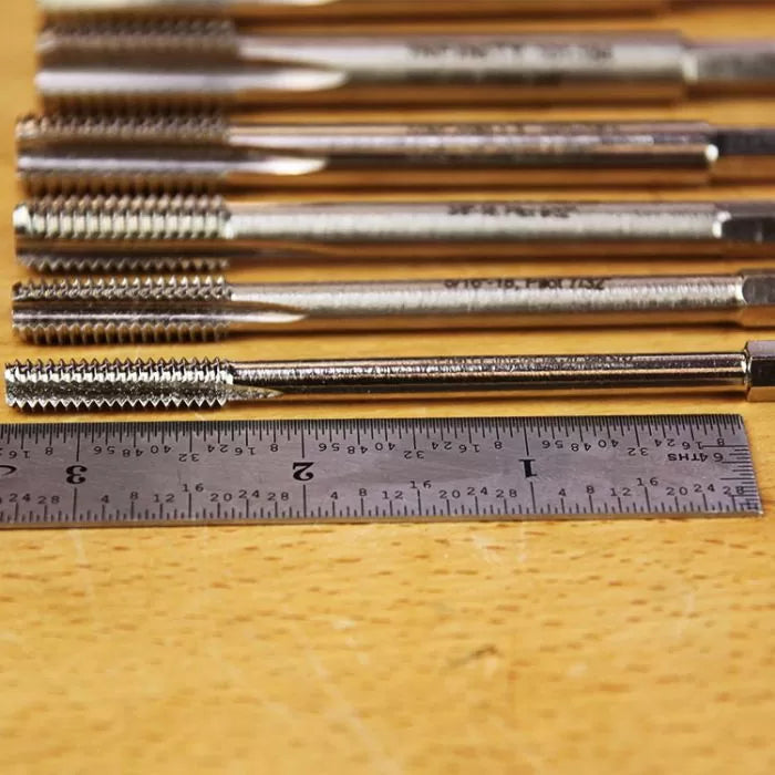 Thread cutting taps lined up next to a metal ruler on a woodworking table