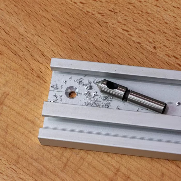 Metal T-track with countersink drill bit and aluminum shavings on a wood workbench.
