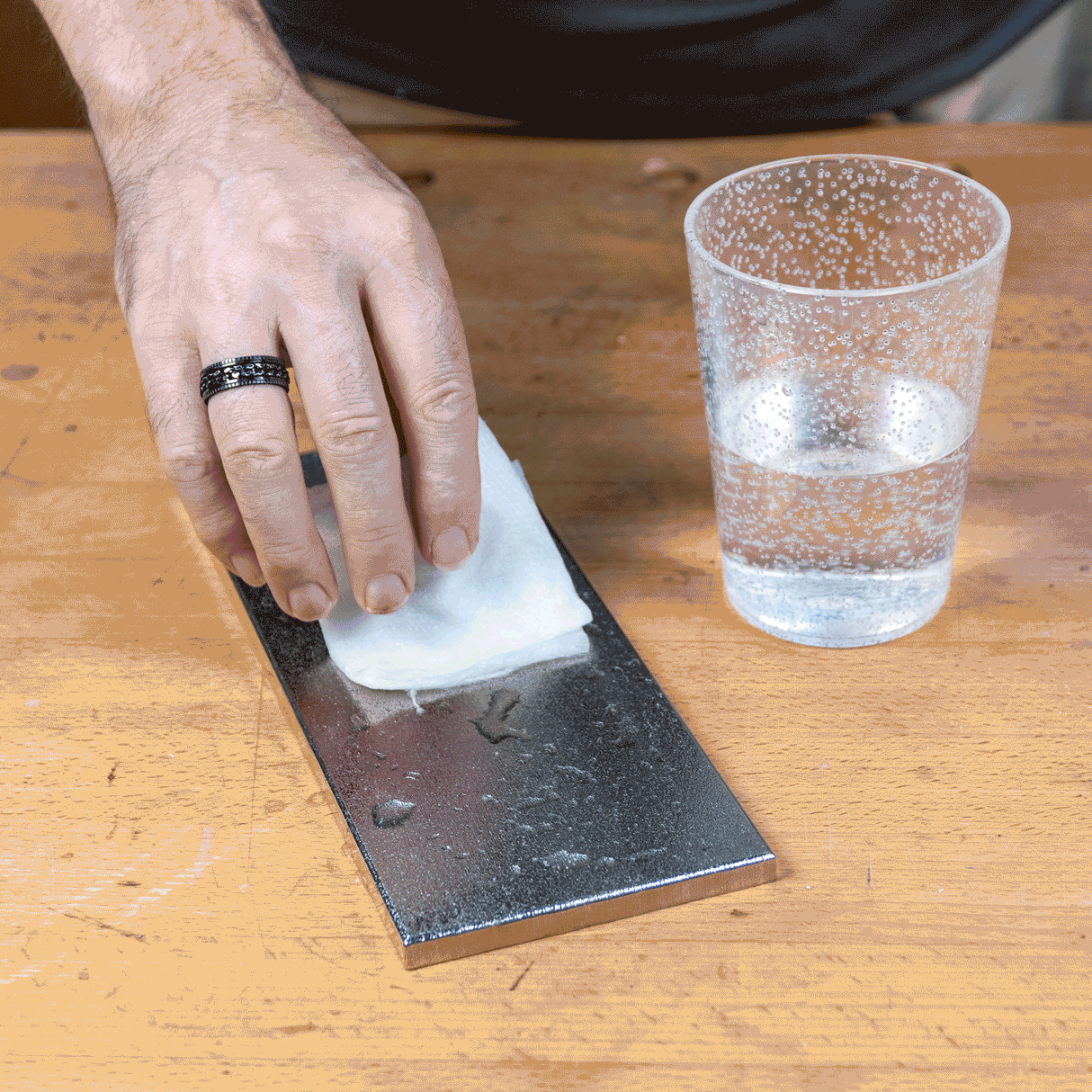 Hand wiping sharpening stone with damp cloth on woodworking bench, water glass nearby