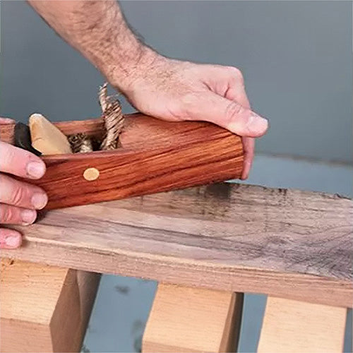 Hand using a wooden hand plane to smooth a wood board on a workbench in a woodworking setting
