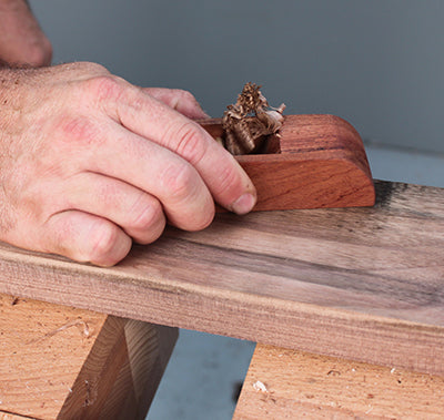 Hand using a wooden hand plane to smooth a hardwood board on a woodworking bench.