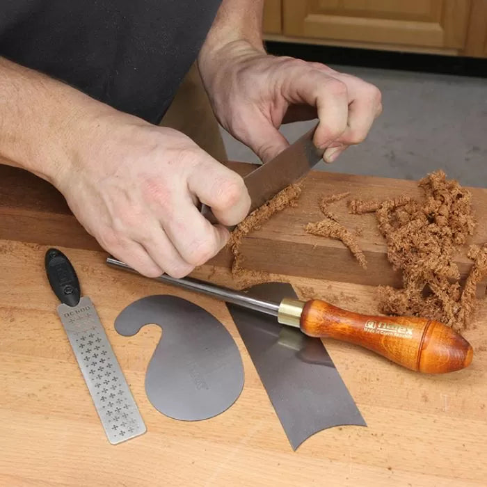 Woodworker using hand scraper on wood block, with cabinet scraper set and sharpening tools on bench.