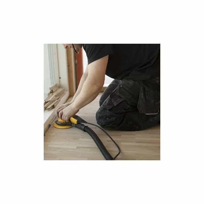 Person using a handheld electric sander on wood flooring near a window, dust hose attached