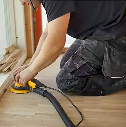 Woodworker using an electric sander on hardwood flooring near a window