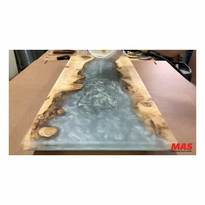 Pouring clear epoxy resin into a live edge wood slab river table on a workshop bench