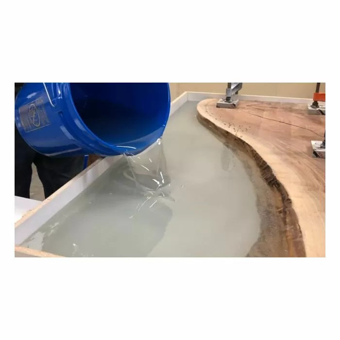 Pouring clear epoxy resin from a blue bucket into a wooden river table mold in a woodworking shop