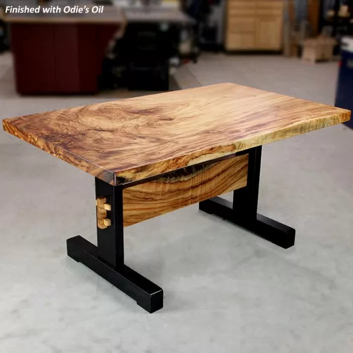 Live edge hardwood table with black metal base, smooth finish, in woodworking shop setting.