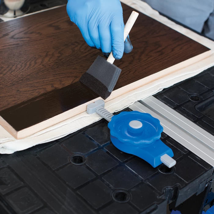 Hand with blue glove using foam brush to stain wooden board on a workbench with a clamp