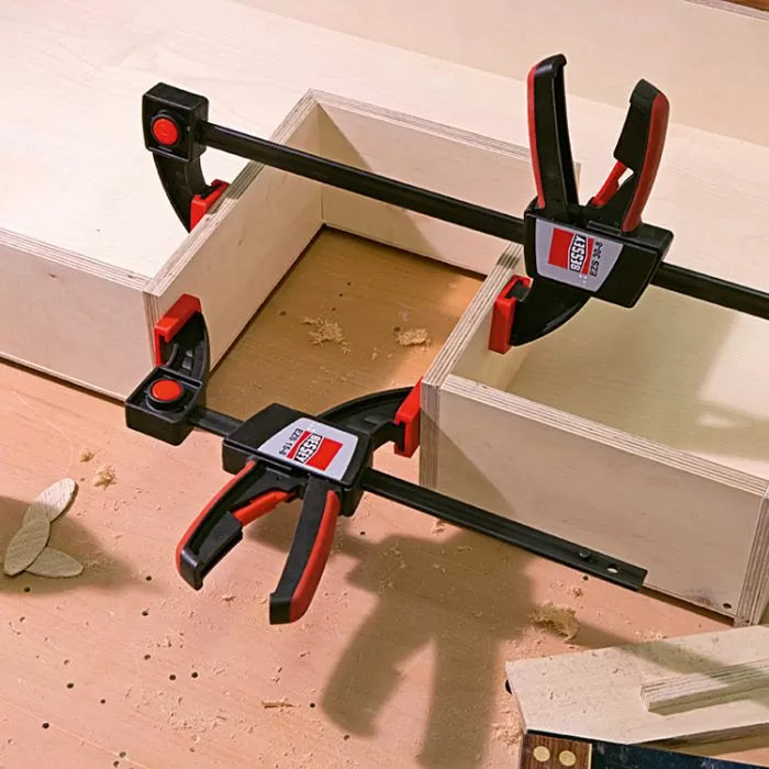 Two quick-grip woodworking clamps securing plywood pieces on a workbench with wood shavings.
