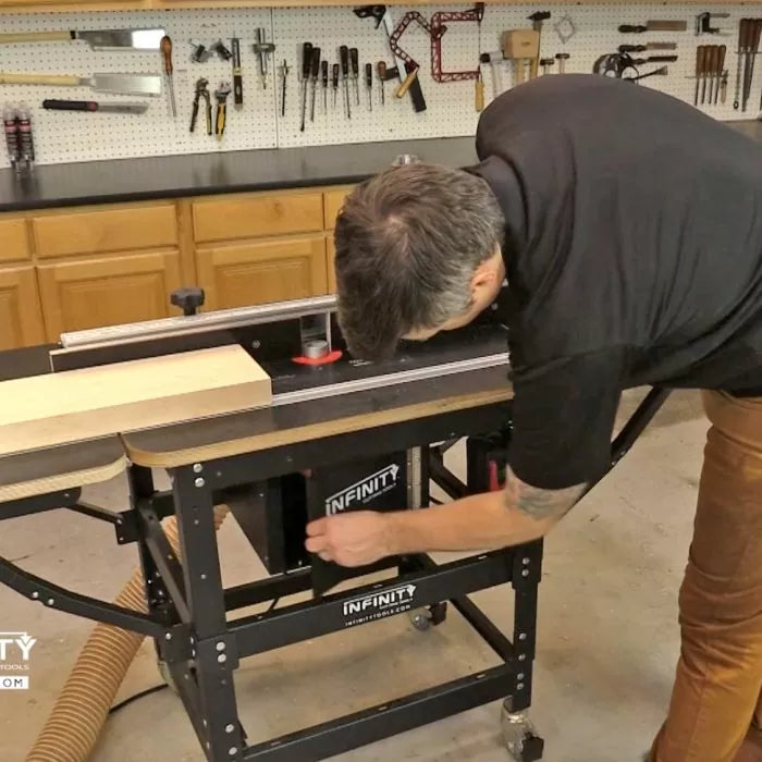 Man adjusting router table in woodworking shop with tools on pegboard wall and wood plank on table.