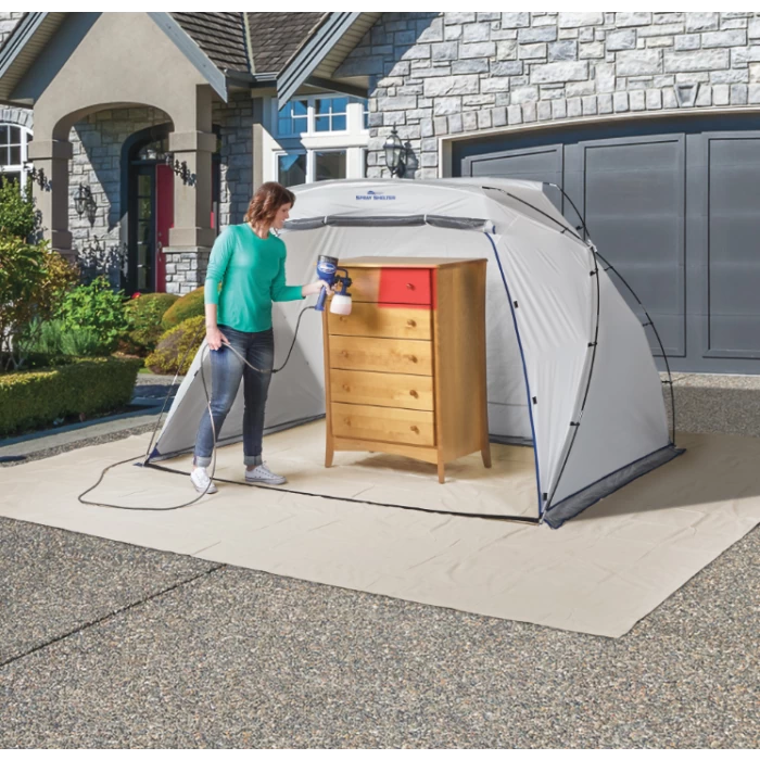 Woman using paint sprayer on wooden dresser inside portable spray shelter outdoors