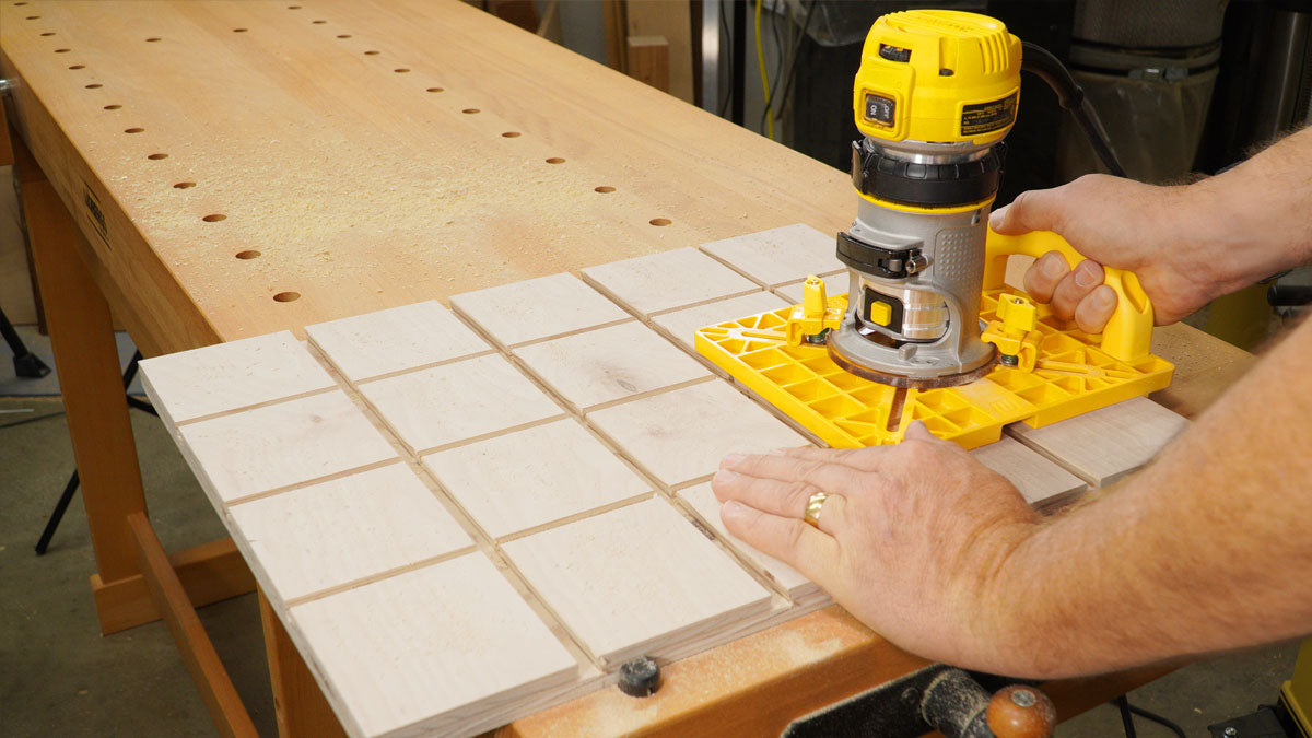 Woodworker using a compact router with a yellow jig to cut grooves in a wood panel on a workbench.