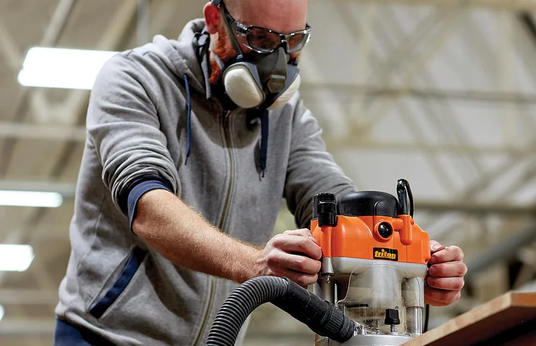 Woodworker using a Triton router on a workbench while wearing safety gear in a workshop.