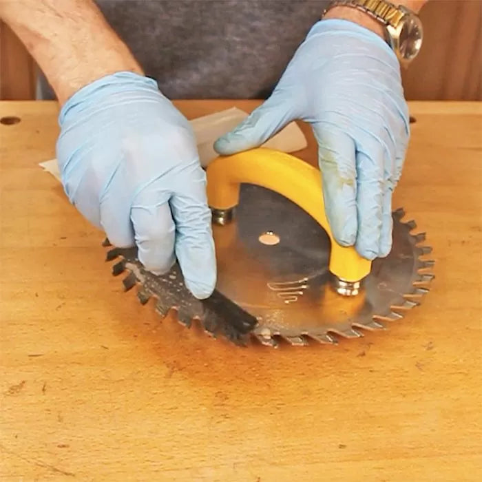 Person cleaning a circular saw blade with a handle and brush on a woodworking bench