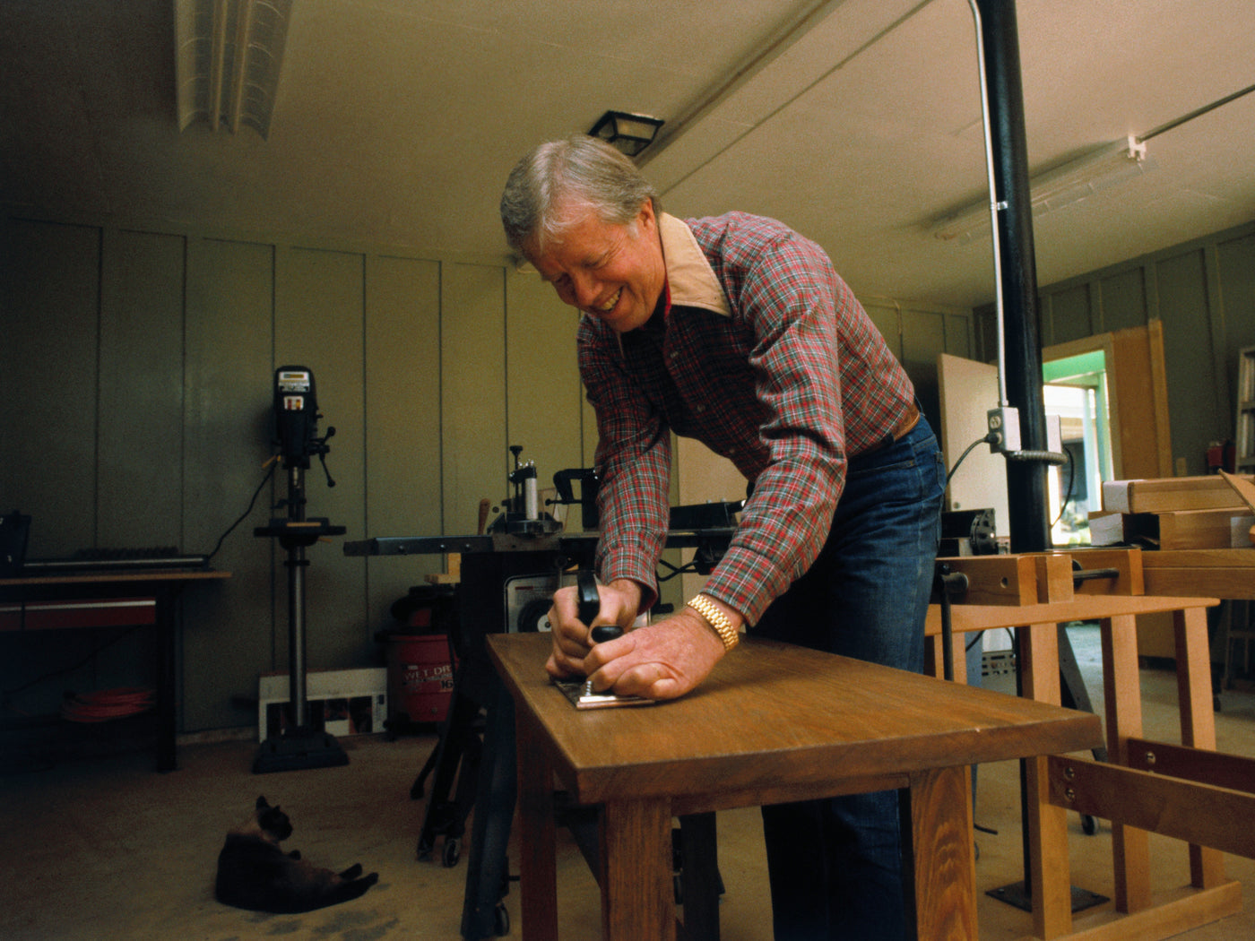 Man woodworking in a workshop, using hand tool on wooden table, surrounded by tools