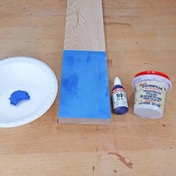 Blue wood filler and pigment with container, wooden board, and pigment bottle on workbench