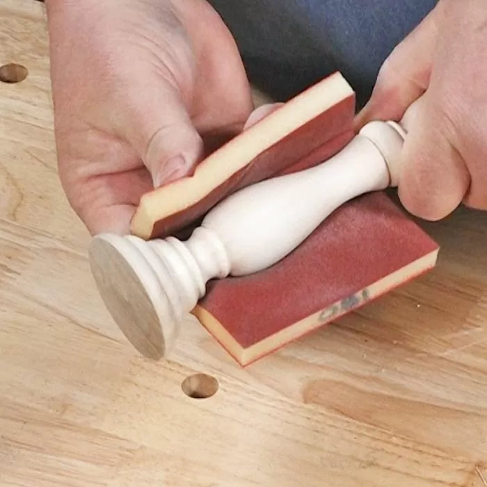 Hand sanding a turned wood spindle with flexible sanding sponge on a workbench