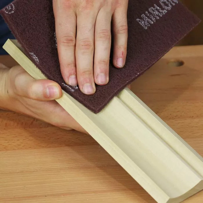 Hand using maroon abrasive pad to sand wooden trim on a workbench