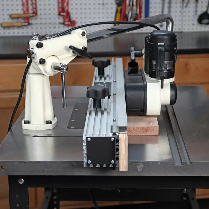Woodworking router table setup with a hold-down featherboard and fence, workshop background