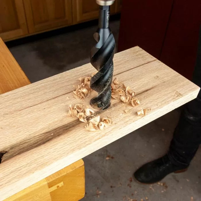 Large wood drill bit creating clean hole in hardwood board with fresh wood shavings