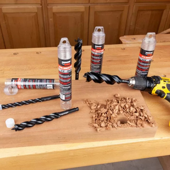 Large wood twist drill bits on workbench with drill, wood chips, and drilled board