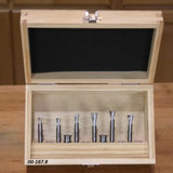 Woodworking router bit set in wooden case on workbench, seven metal bits displayed.