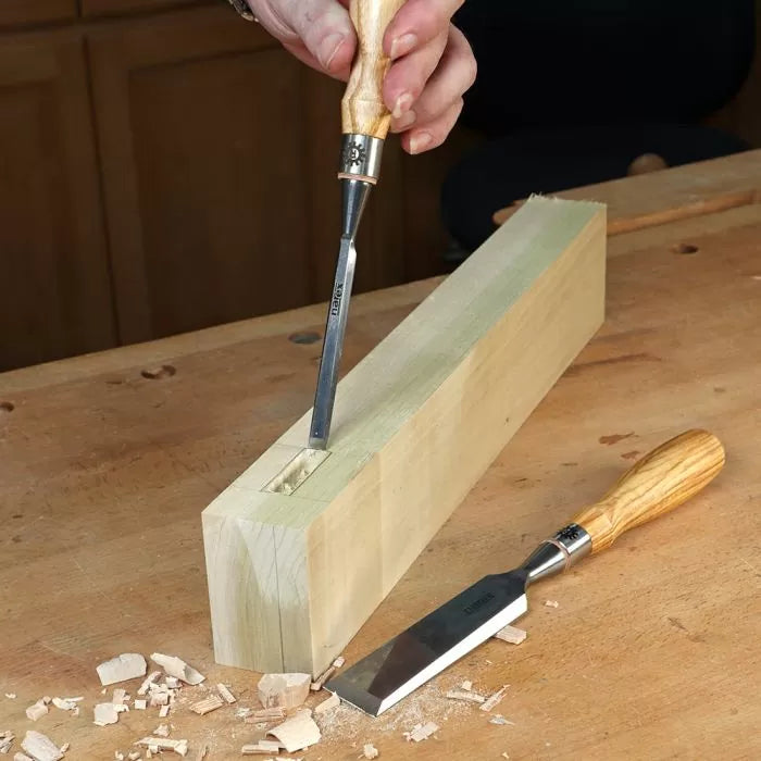 Woodworker using chisel to carve mortise in wooden block on workbench, woodworking tools visible.