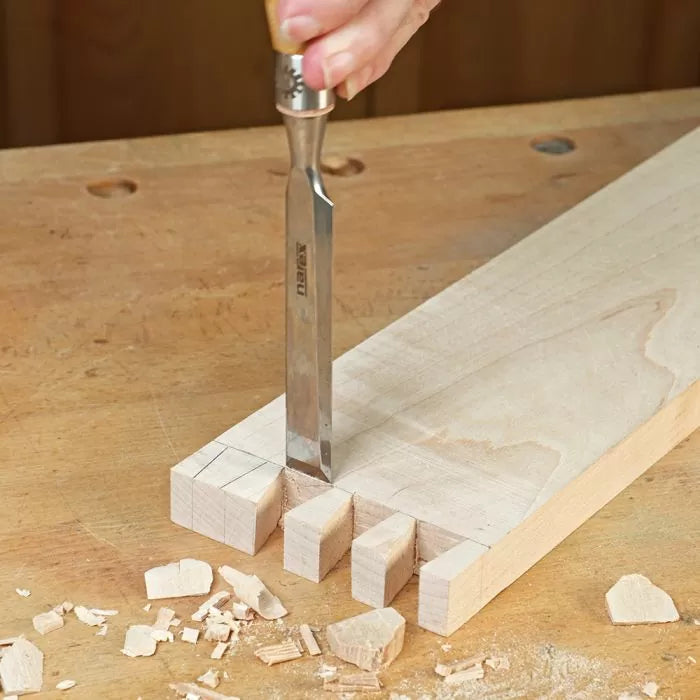 Hand using chisel to cut dovetail joints in wood on a workbench, woodworking tools visible.