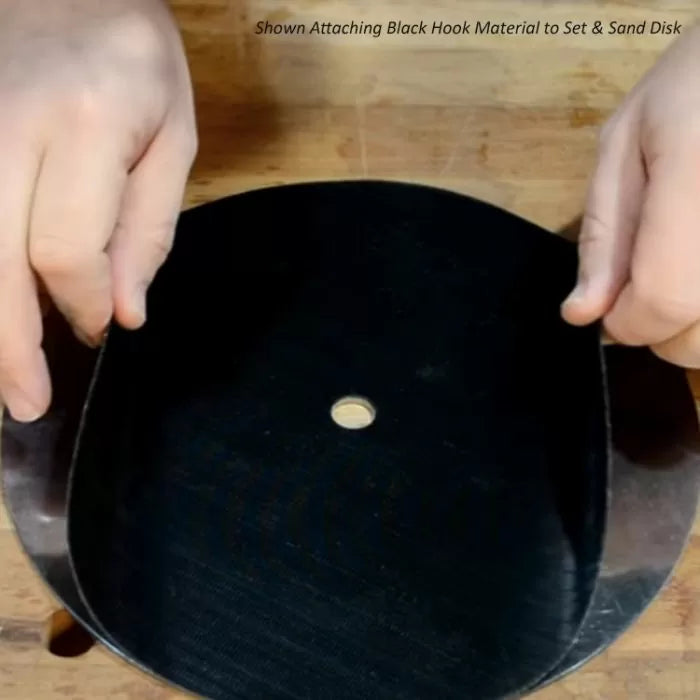 Hands attaching black hook material to round sanding disk on wood surface, woodworking tool