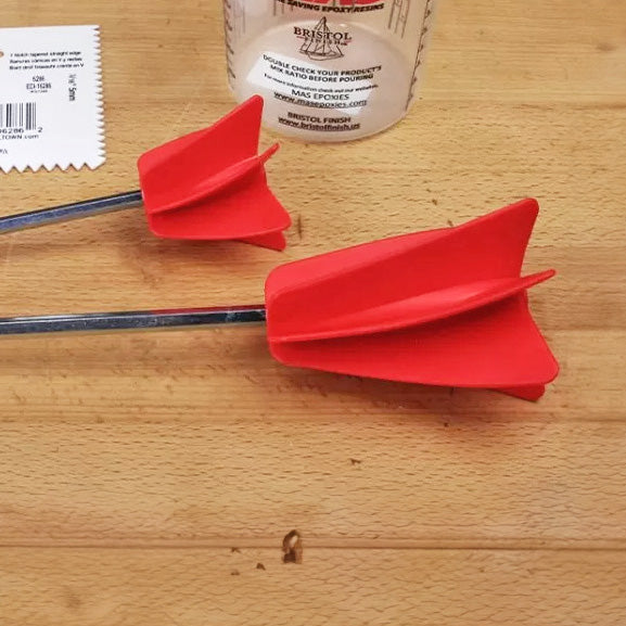 Two red epoxy mixing paddles on a wooden workbench with a measuring cup nearby.