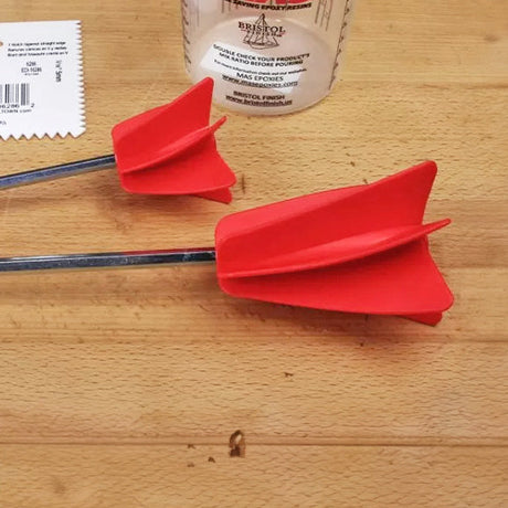 Two red epoxy mixing paddles on a wooden workbench with a measuring cup nearby.