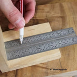 Hand using a pen and 6-inch stainless steel marking rule for precise woodworking measurement on wood.