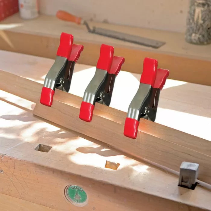 Three red spring clamps securing wood on a workbench in a woodworking shop