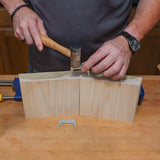 Woodworker using a pinch dog clamp and mallet to join two wood pieces on a workbench.