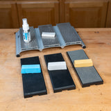 Woodworking sharpening system with three honing stones, abrasive compounds, spray bottle, and base on a workbench.