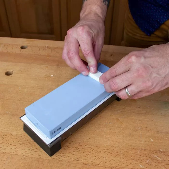 Hands using a blue 1000 grit sharpening stone on a wooden workbench