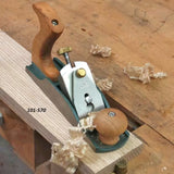 Bench hand plane smoothing wood board on workbench with wood shavings, labeled 101-570.