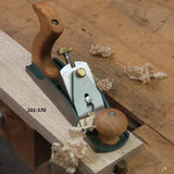 Bench hand plane smoothing wood board on workbench, labeled 101-570, with wood shavings.