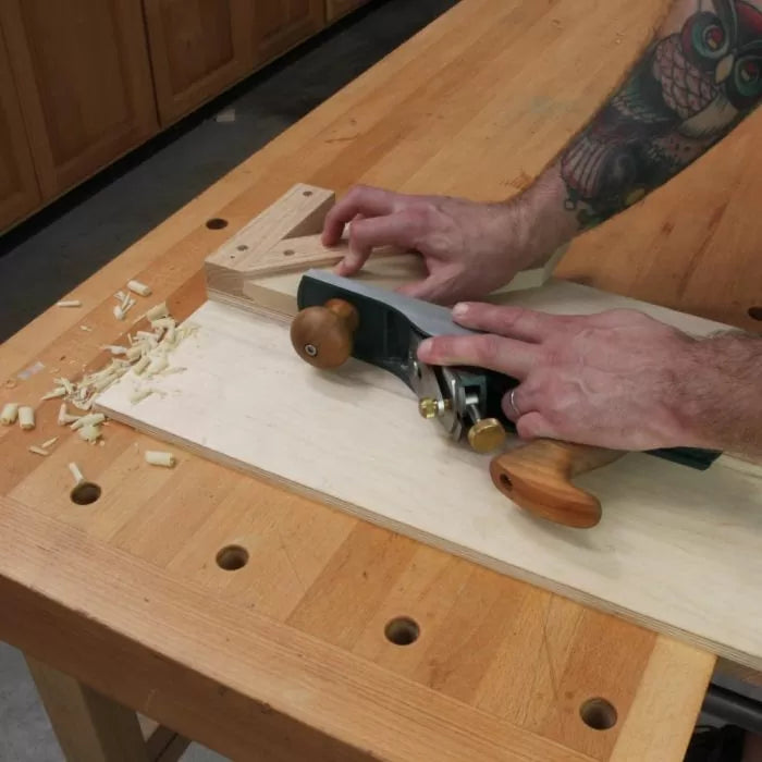 Person using a hand plane on a woodworking shooting board with wood shavings on bench