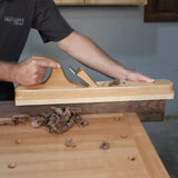 Woodworker using a wooden jointer hand plane to smooth hardwood edge on workbench.