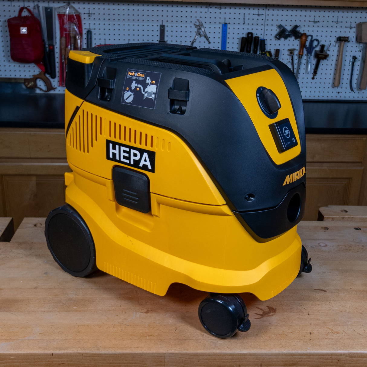 A yellow and black HEPA vacuum cleaner with caster wheels, positioned on a wooden workbench in a workshop setting.