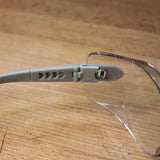 Clear safety glasses with gray adjustable arm on a wooden surface, woodworking safety gear