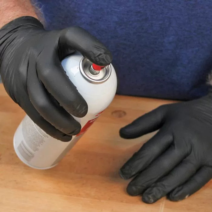 Person wearing black gloves holding adhesive spray can on woodworking table