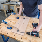 Woodworker using clamps and cordless drill to assemble wood on workbench in workshop