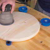 Hand sanding round wooden board held with blue woodworking bench clamps