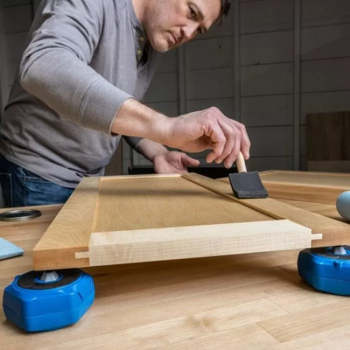 Man applying finish with foam brush to wooden cabinet door on workshop table, woodworking tools nearby