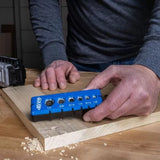 Woodworker using a blue drill bit gauge tool on a wooden board in a workshop