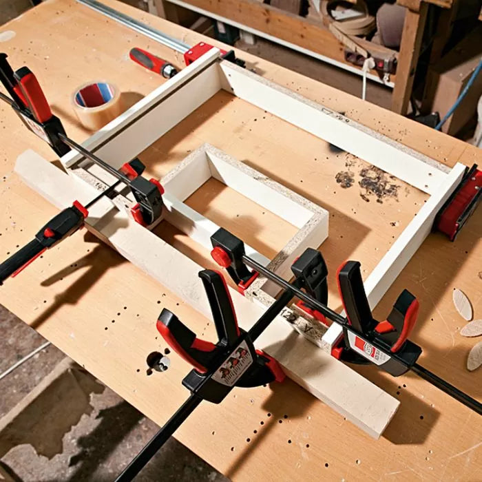 Woodworking clamps securing white boards together on a workbench in a workshop setting.