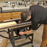Man adjusting router table in woodworking shop with tools on pegboard wall and wood plank on table.